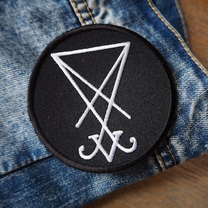 May include: A black and white embroidered patch with a white symbol on a black circle. The symbol is a triangle with a line through it and two curved lines at the bottom.