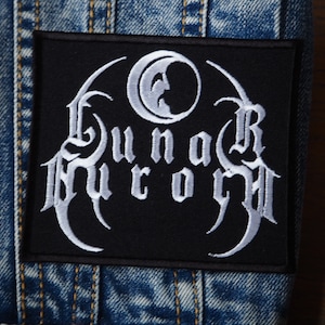 Puede incluir: Parche bordado negro con una luna creciente blanca y el texto "Lunar Aurora" en una fuente gótica.