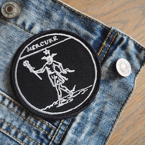 May include: Black and white embroidered patch with the word "MERCURIE" and a stylized image of a figure with a staff. The patch is sewn onto a blue denim jacket.