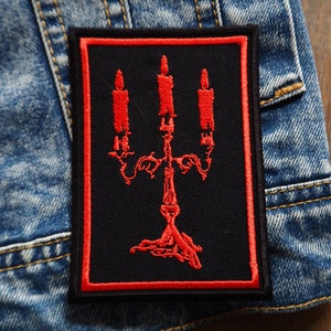 Puede incluir: Parche bordado rojo con fondo negro que presenta un candelabro con tres velas. El parche tiene un borde rojo.