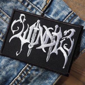 Peut inclure: Écusson brodé noir avec le mot "Swords" en lettres blanches. Les lettres sont stylisées avec une police gothique et comprennent deux épées.