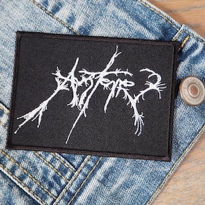 Puede incluir: Parche negro con texto bordado en blanco "Aether" en una chaqueta vaquera.