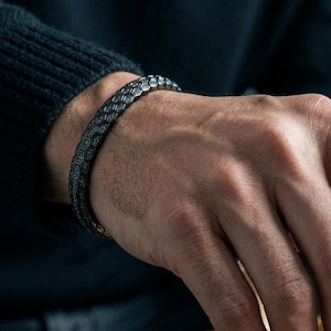 May include: A dark, patterned bracelet adorns a wrist, set against a dark background. A hand moves a dark chess king on a wooden chessboard. The chess pieces are a mix of dark and light wood tones, creating a classic game scene.