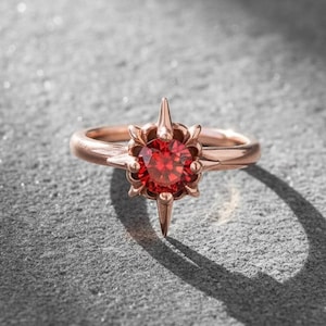 May include: Rose gold ring with a central, round, red gemstone. The ring has a starburst design around the gemstone and a pointed element extending from the bottom. The ring is set against a textured grey surface.