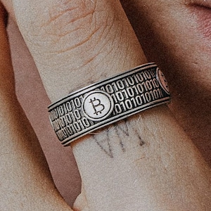 May include: Silver ring with a Bitcoin symbol and binary code design. The ring is worn on a finger, with the Bitcoin symbol at the centre. The ring's design includes the Bitcoin symbol and binary code. The ring is a piece of jewellery.