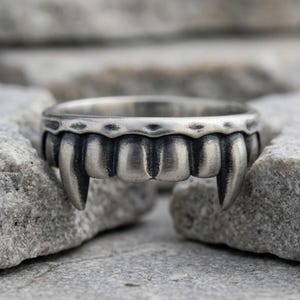 May include: A silver-toned metal ring featuring a sculpted design of a row of teeth with two prominent fangs on either side. The surface of the ring has a darkened, oxidized finish in the recessed areas to highlight the individual tooth shapes.