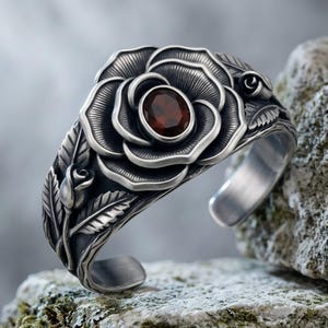 May include: A silver cuff bracelet featuring a large, detailed rose motif with an oval-shaped, deep red gemstone set in the center. The band is decorated with sculpted leaves and smaller rose buds, showcasing an oxidized finish for depth.