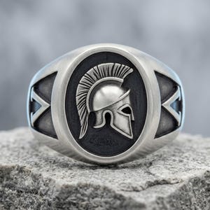 May include: A silver-toned ring featuring a central oval inlay with a raised, detailed relief of a Spartan helmet. The helmet is depicted in profile with a prominent crest. The band has a brushed finish with dark, recessed geometric accents.
