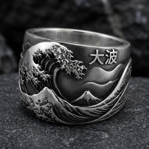 May include: A wide silver ring featuring a detailed relief of a crashing ocean wave and a mountain peak. The metal has an oxidized finish to highlight the textures. The Japanese characters 大波 are engraved on the upper right side of the band.