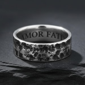 May include: A silver ring with a textured, hammered finish. The inside of the ring features the engraved words "AMOR FATI". The ring is set against a dark background.