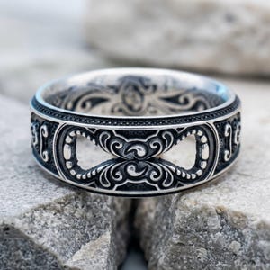 May include: A silver ring featuring an intricate, oxidized relief pattern with repeating scrollwork and cutout infinity-like shapes. The metal surface displays a high-contrast finish, highlighting the detailed engravings along the band's exterior.