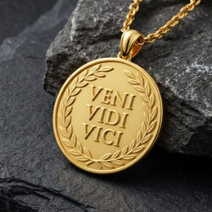 Puede incluir: Un collar con colgante dorado con un medallón redondo. El medallón presenta la frase latina "VENI VIDI VICI" dentro de un diseño de corona de laurel. La cadena y el colgante son de metal brillante y dorado.