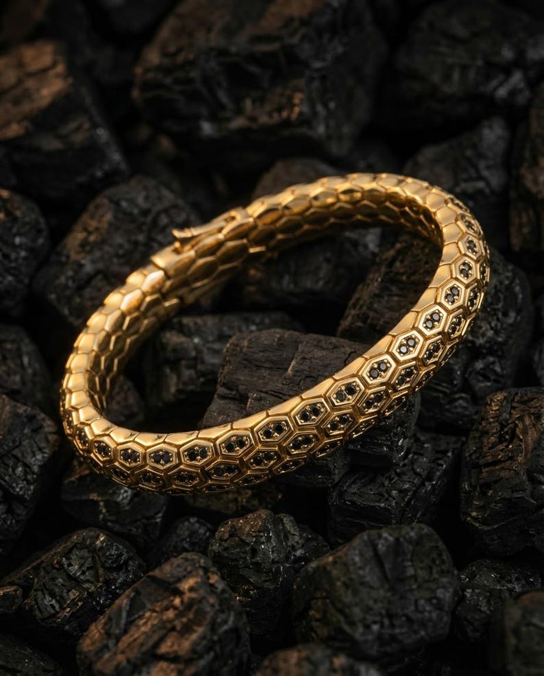 Puede incluir: Un brazalete dorado con un dise&ntilde;o de panal, con peque&ntilde;os detalles negros en cada celda. El brazalete se encuentra sobre un fondo de piedras oscuras y texturizadas, creando un contraste entre el metal brillante y la superficie rugosa.