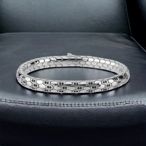 Pode incluir: Uma pulseira prateada com um design de favo de mel. A pulseira é feita de elos hexagonais interligados, com um padrão repetido de pequenos detalhes pretos dentro de cada hexágono. A pulseira é exibida em uma superfície preta.