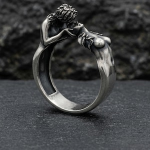 Può includere: Un anello d'argento con un design scolpito di due figure che si abbracciano. L'anello raffigura un uomo e una donna in una posa romantica, con dettagli nei capelli e nei corpi. L'anello è realizzato in un materiale metallico lucido.