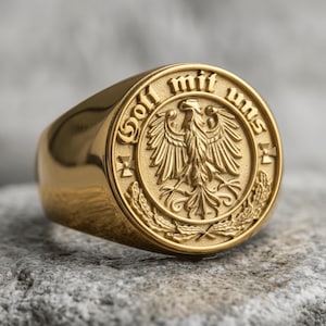 May include: A gold-toned signet ring featuring a circular face with an embossed eagle emblem at the center. The text "Gott mit uns" is engraved along the upper edge of the circular border, flanked by small cross symbols and laurel leaf motifs.