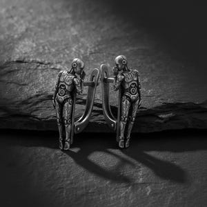 May include: A pair of silver-toned robot-shaped earrings. Each earring features a detailed robot figure with intricate mechanical designs. The robots are standing, with one hand raised to their face. The earrings are displayed on a dark, textured surface.
