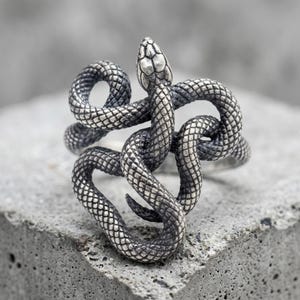 May include: A silver-toned ring shaped like a coiled snake with detailed scales. The metal has an oxidised finish that highlights the texture of the snake's body, which wraps around to form the band. The snake's head is positioned at the top.