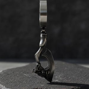 May include: A silver-colored pendant in the shape of a stylized axe. The pendant has intricate details, including a textured handle and a blade with a detailed design. The pendant is suspended from a silver ring.