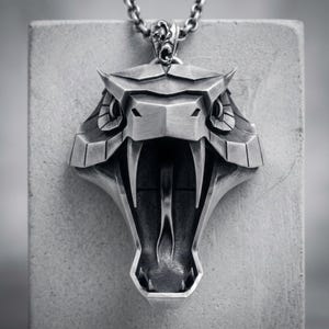 Pode incluir: Um pingente de metal prateado em forma de cabeça de cobra estilizada com a boca aberta, presas proeminentes e língua bífida. O pingente apresenta detalhes geométricos angulares e está suspenso por uma corrente de elos de metal prateado.