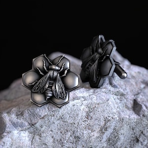 May include: A pair of silver cufflinks featuring a bee on a honeycomb design. The cufflinks are on a gray rock surface.