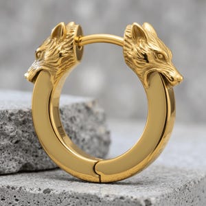 May include: A gold-toned hoop earring featuring two sculpted wolf heads facing each other at the top of the circular band. The metal has a polished, reflective finish with detailed fur textures on the wolf heads and a smooth, curved hoop design.
