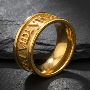 May include: A gold-colored ring with the inscription "Veni Vidi Vici" engraved around the band. The ring has a polished, reflective surface and a classic, unisex design. The ring is displayed on a dark, textured surface.