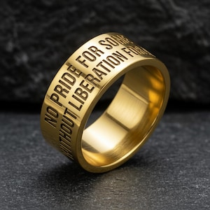 Op de afbeelding: Een goudkleurige ring met de gegraveerde tekst "NO PRIDE FOR SOME WITHOUT LIBERATION FOR ALL". De ring is gemaakt van glanzend metaal en staat tegen een donkere, getextureerde achtergrond.