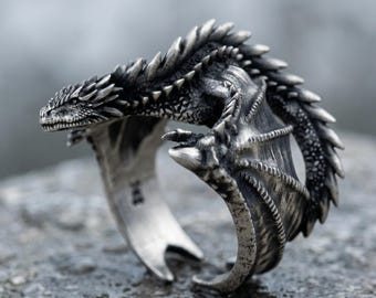 May include: A silver-toned metal ring shaped like a coiled dragon with intricate scales, a textured wing, and a pointed tail. The dragon's head faces forward, and its body forms the band of the ring, featuring a dark, oxidized finish for detail.