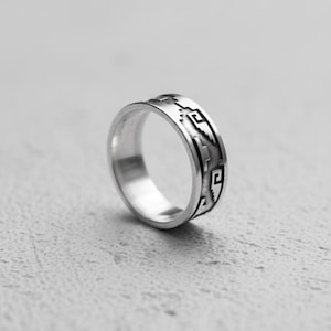 Può includere: Un anello d'argento con un motivo geometrico. L'anello è di forma circolare con una fascia di forme dettagliate e ripetute all'esterno. È posto su uno sfondo grigio chiaro.