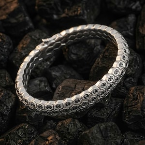 May include: A silver bracelet with a honeycomb pattern, each cell filled with a small black stone. The bracelet is displayed on a bed of dark, textured stones. The design is intricate and the contrast between the silver and black is striking. A stylish piece of jewellery.