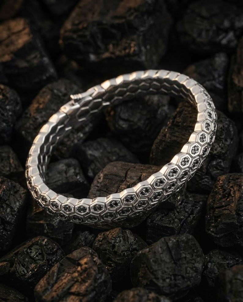 Puede incluir: Una pulsera plateada con un dise&ntilde;o de panal, con peque&ntilde;os detalles negros en cada celda. La pulsera se encuentra sobre un fondo de piedras oscuras y texturizadas. El dise&ntilde;o es intrincado y el contraste plata/negro es llamativo. Una joya elegante.