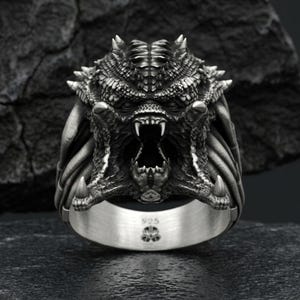 Peut inclure: Bague en argent représentant une tête de Predator détaillée, avec une bouche ouverte, des dents acérées et une peau texturée. La bague est marquée "925" et d'un petit emblème sur le bandeau. Bijou de style fantastique.