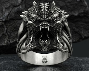 May include: A silver ring with a detailed Predator head design. The ring features an open mouth with sharp teeth and textured skin. The band is marked with "925" and a small emblem. The ring is a striking piece of jewellery.