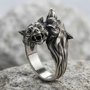 May include: A silver-toned metal ring featuring a detailed sculptural design of two wolf heads. One wolf head is depicted with an open mouth showing teeth, while the other is positioned behind it with its head tilted upward in a howling posture.