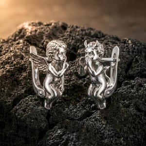 May include: A pair of silver-toned earrings, one depicting an angel with wings and the other a devil with horns. The earrings are detailed with intricate designs and are set against a dark, textured background.