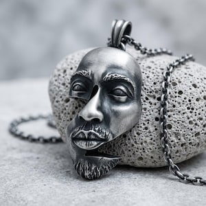 Puede incluir: Un colgante de metal plateado con forma de rostro humano con un diseño segmentado y en capas, que presenta ojos, nariz, labios y barba detallados. El colgante está unido a un collar de cadena de eslabones de metal plateado a juego.