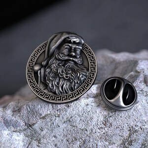 Puede incluir: Un pin de solapa circular de metal que presenta un retrato en relieve detallado de una figura barbuda con sombrero. El borde del pin está decorado con un patrón geométrico de greca. Un cierre de metal separado es visible junto al pin.