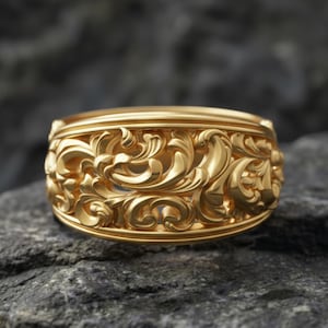 Puede incluir: Un anillo dorado con un intrincado diseño floral. El anillo presenta tallas detalladas de hojas y vides arremolinadas, creando una superficie texturizada. El anillo está sobre un fondo oscuro y texturizado.