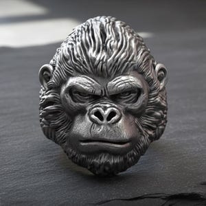 May include: A detailed silver-colored ring shaped like a gorilla's head. The ring features intricate detailing of the gorilla's fur and facial features, including a stern expression. The ring is displayed on a dark, textured surface.