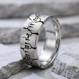 Puede incluir: Un anillo de metal plateado con acabado cepillado, que presenta una grieta vertical prominente en el centro. La superficie está grabada con símbolos lineales oscuros que se asemejan a runas antiguas, situados dentro de la fisura.