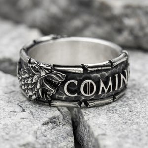 May include: A silver-toned metal ring featuring a detailed relief of a wolf head on one side and the text COMIN with a sword icon replacing the letter I. The band has a dark, hammered texture background and a polished, raised metallic finish.