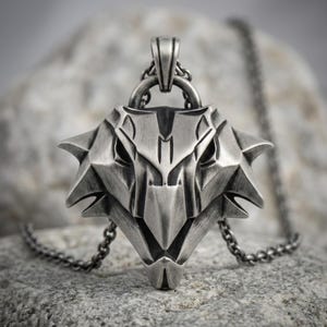 May include: A silver-toned metal pendant shaped like a stylized wolf head with sharp, angular features and a dark, weathered finish. The pendant hangs from a matching silver-toned metal chain, featuring a prominent bail at the top for suspension.