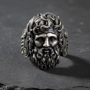 May include: A silver ring featuring a detailed depiction of a bearded man's face, possibly a Greek god. The ring has intricate curly hair and beard details, with decorative elements on the sides. The ring is set against a dark, textured background.
