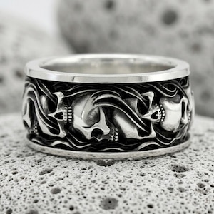 May include: A wide silver band ring featuring a continuous relief pattern of multiple skulls integrated into swirling, dark-oxidized wave designs. The polished silver edges frame the detailed, recessed sculptural work around the entire circumference.
