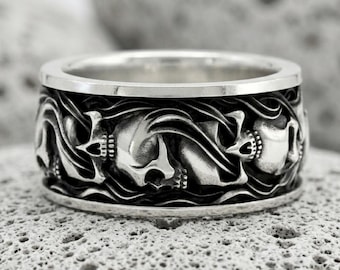 May include: A wide silver band ring featuring a continuous relief pattern of multiple skulls integrated into swirling, dark-oxidized wave designs. The polished silver edges frame the detailed, recessed sculptural work around the entire circumference.