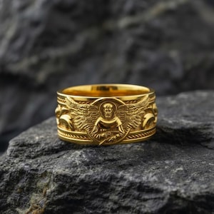 Könnte beinhalten: Ein goldfarbener Ring mit einem aufwendigen Design, das eine geflügelte Figur zeigt. Der Ring hat ein breites Band mit detaillierten Reliefschnitzereien. Der Ring wird auf einer dunklen, strukturierten Oberfläche präsentiert.