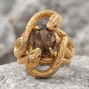 May include: A gold ring featuring two intertwined snakes with textured scales and small red gemstone eyes. The snakes encircle a central oval-cut smoky brown gemstone. The metal has a polished finish, and the design creates a continuous loop.