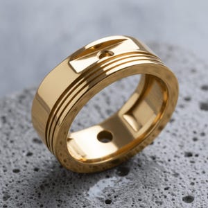 Puede incluir: Un anillo de tono dorado pulido que presenta una banda ancha con acabado liso, tres ranuras horizontales en el lateral y una pequeña hendidura circular en la superficie superior. El anillo es hueco con un orificio visible en la pared interior.