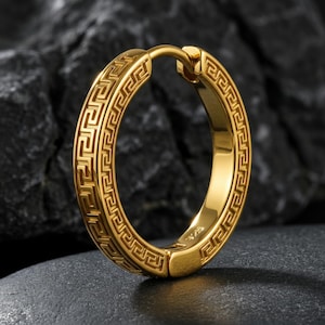 May include: A gold-coloured hoop earring featuring a Greek key design. The earring is oval and has a hinged clasp. The earring is shown against a dark, textured backdrop.